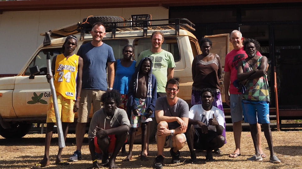 A group of smiling Indigenous and non-Indigenous people stands smiling in front of a dusty 4 wheel drive in Mandjawuy, NT