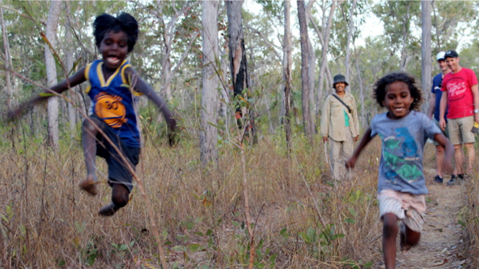 2 Indigenous children run and jump through the bush while 3 smiling adults watch on