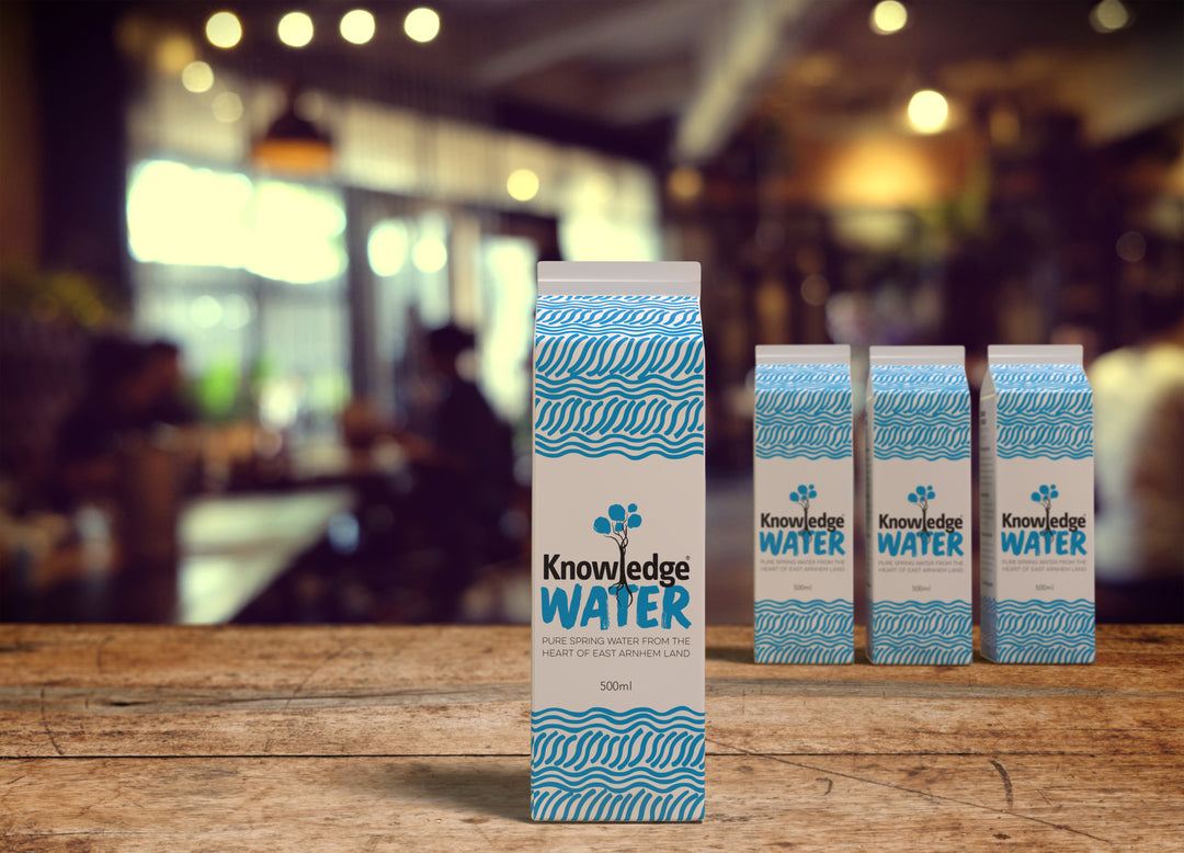 4 forward-facing 500ml cartons of Knowledge Water on a timber table in front of a blurry cafe background
