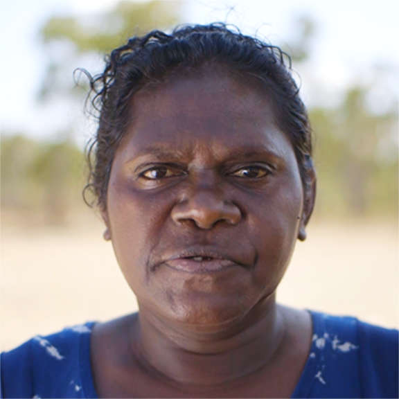 A proud Indigenous woman looks at the camera in front of a blurred bush background