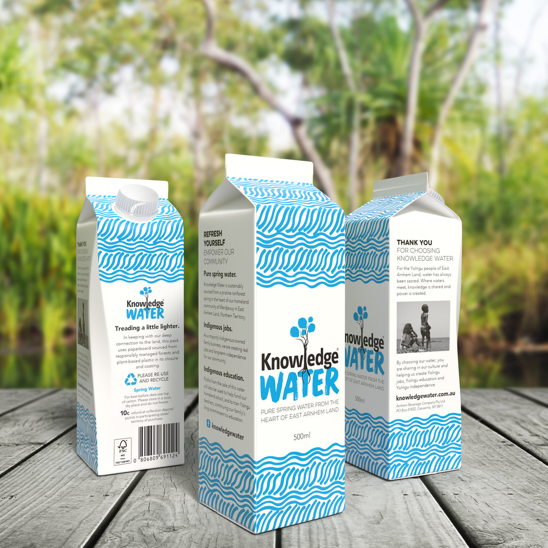 Three cartons of Knowledge Water on a timber deck in front of a rainforest spring at the Manjawuy Indigenous Community